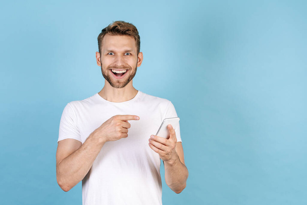 Concept on new online app. Happy and excited adult man pointing with finger on modern smartphone, standing isolated on blue copy space background, wearing in white t-shirt - Φωτογραφία, εικόνα