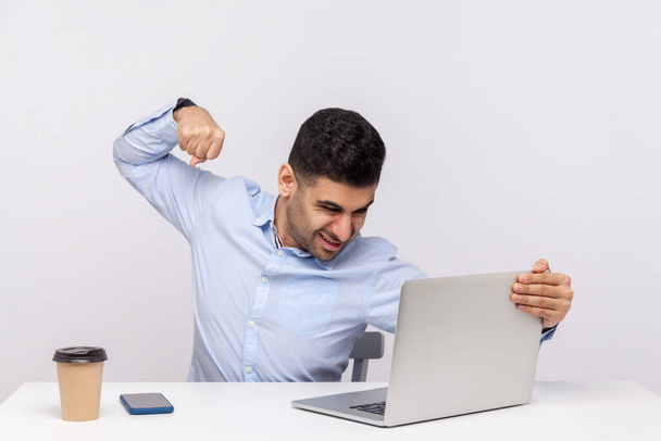 Angry businessman punching laptop screen, looking with furious mad expression, boxing threatening to hit while having online conversation, video call. indoor studio shot isolated on white background - 写真・画像