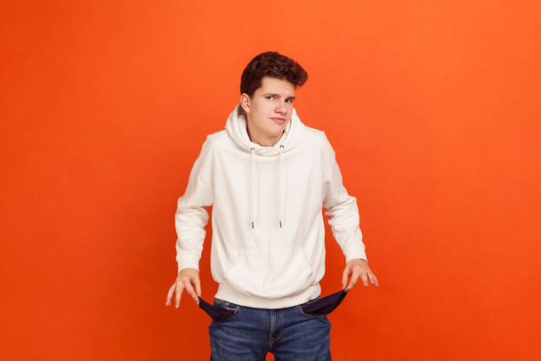 I have no money, Poor teenager in casual sweatshirt with hood demonstrating empty pockets of his modern denim pants, low state stipend, bankruptcy. Indoor studio shot isolated on orange background - Фото, изображение