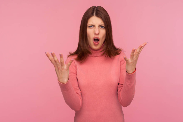 Nervous disappointed brunette woman in pink sweater spreading hands and shouting asking what do you want from me? Indoor studio shot isolated on pink background - Photo, Image