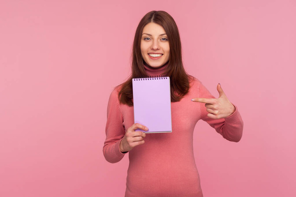 Positive smiling woman pointing finger showing notebook with blank space for commercial text, advertising area on paper for plans. Indoor studio shot isolated on pink background - Fotó, kép