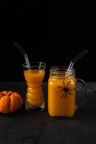 Non-alcoholic pumpkin mocktail in drinking glass and square jar with glass straws and whole orange pumpkin on black textured surface. Halloween spider decor on jar. Healthy eating concept. Copy space. - Fotografie, Obrázek