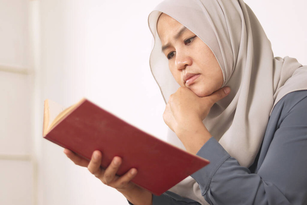 Beautiful Asian muslim woman reading book while sitting on sofa, girl enjoys her time by doing leisure activity, happy cheerful expression - Photo, Image