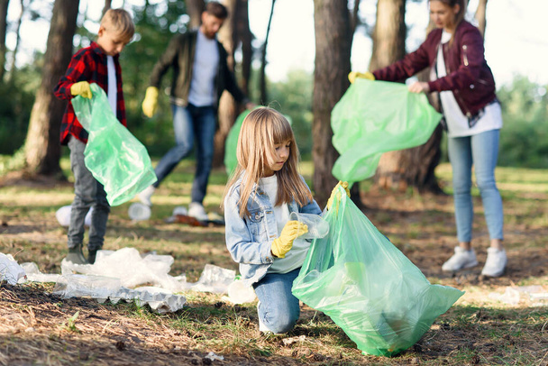 Motivated beautiful girl picking up rubbish at the backdrop of multi aged volunteers at the park. Anti pollution concept - Фото, зображення