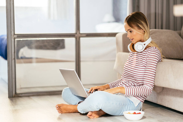 Smiling young woman with headphones and laptop on the sofa - Фото, изображение