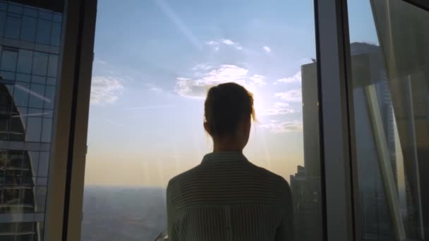 Zamyślona kobieta patrząca na pejzaż miasta przez okno wieżowca - widok z tyłu - Materiał filmowy, wideo