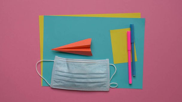 Face mask on top of coloring pens and papers with paper plane. Pink background. - Foto, Imagen