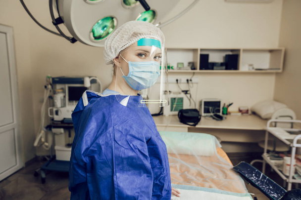 Una joven cirujana con una máscara protectora, gafas y un escudo en el quirófano. Un médico en un hospital durante una pandemia de coronavirus. Operación durante la cuarentena - Foto, Imagen