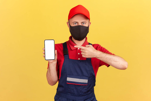 Repairman in blue overalls red t-shirt and hat with protective mask on face pointing finger at smartphone with empty display, online service order app. Indoor studio shot isolated on yellow background - Valokuva, kuva