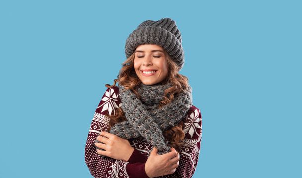 Zimowa moda. Piękna młoda kobieta nosi wełniany sweter, czapkę z dzianiny i szalik na niebieskim tle studio - Zdjęcie, obraz