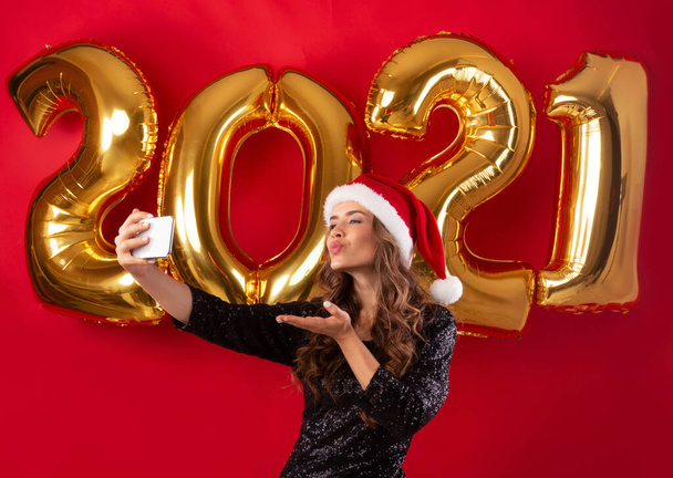 Elegante yong vrouw in Santa hoed en mooie avond jurk nemen selfie op oudejaarsavond 2021, rode studio achtergrond - Foto, afbeelding