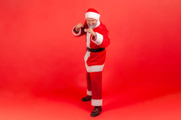 Full length portrait of happy purposeful gray bearded man in red santa claus costume and eyeglasses holding clenched fists up ready to boxing. Indoor studio shot isolated on red background - Zdjęcie, obraz