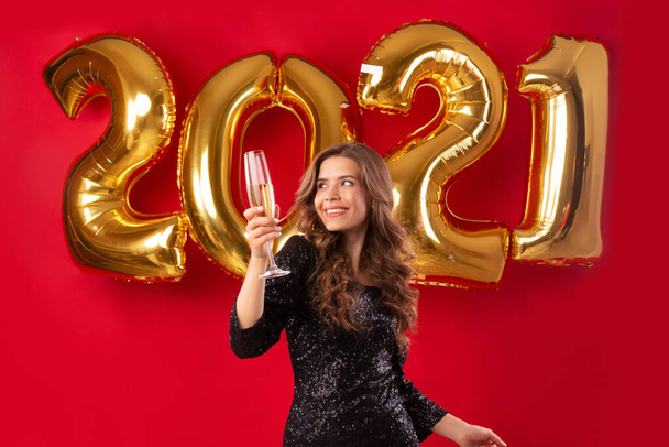 Süße Millennial-Frau trinkt Champagner während der Neujahrsparty 2021 auf rotem Studiohintergrund mit goldenen Luftballons - Foto, Bild