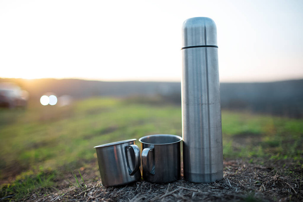 Close-up de termo de aço e duas xícaras de aço inoxidável na grama verde ao pôr do sol. - Foto, Imagem