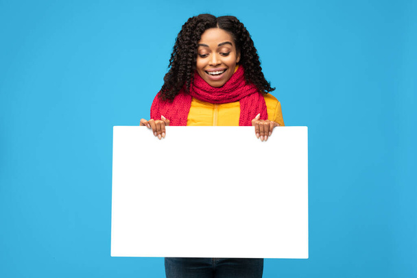 zwart Millennial vrouw houden lege poster staande over blauwe achtergrond - Foto, afbeelding