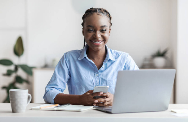 Junge schwarze Büroangestellte sitzt mit Smartphone in der Hand am Arbeitsplatz - Foto, Bild