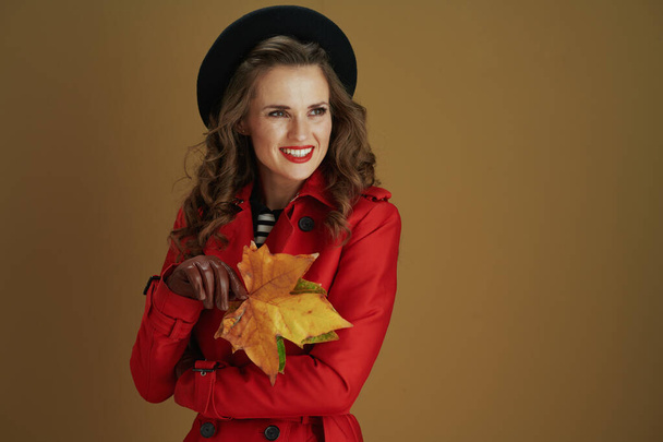 Hello autumn. happy stylish middle aged woman in red coat and black beret with leather gloves and yellow autumn maple leaves looking into the distance isolated on beige. - Photo, image