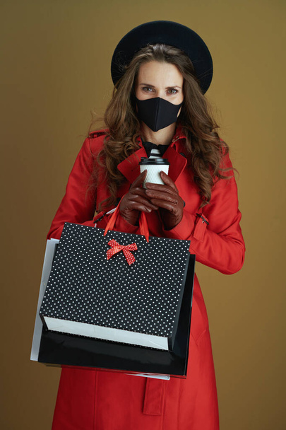 Dobrý den. mladá hospodyňka v červeném kabátě s černou maskou, pohár a papírové nákupní tašky izolované na bronzovém pozadí. - Fotografie, Obrázek
