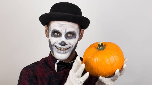 A horrible man in clown make-up holds a pumpkin, symbol of Halloween. A scary clown looks at the camera, holds a pumpkin in his hands and threatens her with a knife. Scary grimaces of a clown looking at the camera. Halloween costume. - Foto, Imagem