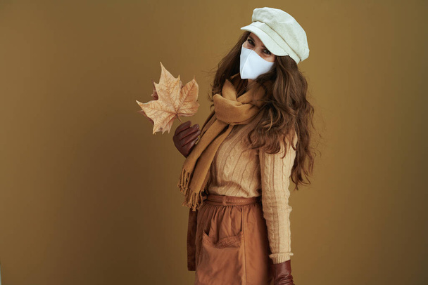 Life during coronavirus pandemic. smiling stylish woman in sweater with white medical mask and yellow autumn maple leaves against beige background. - Fotoğraf, Görsel