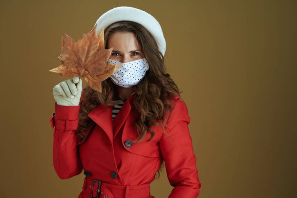 Life during covid-19 pandemic. smiling modern woman in red coat with yellow autumn maple leaf against brown background. - Фото, изображение