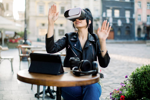 Tecnología digital, realidad virtual. Feliz joven morena positiva, moviendo las manos mientras intenta gafas VR auriculares durante el trabajo o estudio en el café de la ciudad al aire libre - Foto, imagen