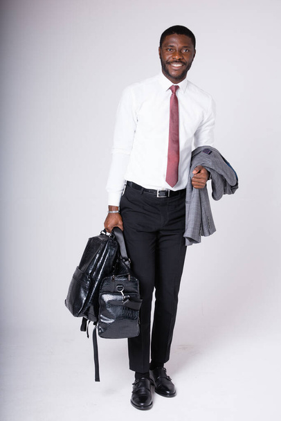 An African American man stands with a large business travel bag and a jacket in his hand on a gray background. Business and work travel - Valokuva, kuva