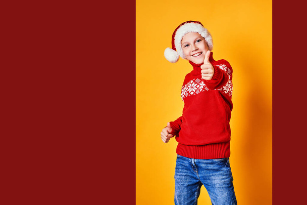 Boy in Santa hat and red christmas design sweater having fun and showing thumb up on Christmas day against yellow background - Valokuva, kuva