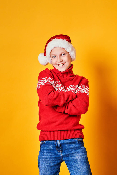 Boy in Santa hat and red sweater posing on Christmas day against yellow background - Фото, зображення