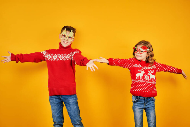 Children in warm red Christmas sweaters and decorated glasses looking at camera on yellow background - Photo, Image