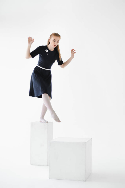 Young woman balances on high cubes on a white background. Studio portrait of a girl in a dress and white tights barefoot stepping from one support to another. Fear of taking a step forward, caution. - Foto, Imagem