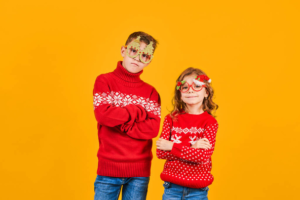 Children in warm red Christmas sweaters and decorated glasses looking at camera on yellow background - Foto, Imagen