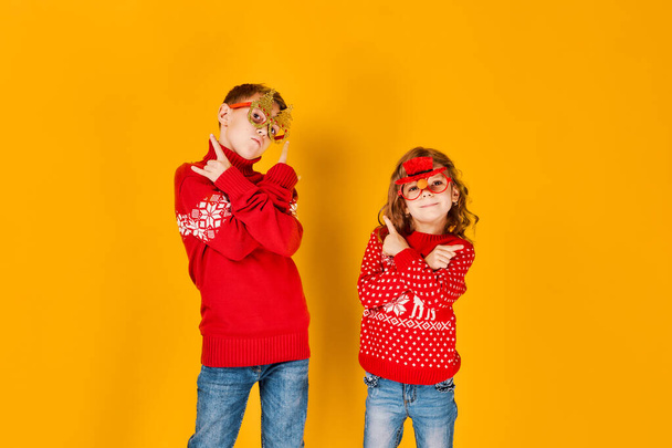 Children in warm red Christmas sweaters and decorated glasses looking at camera on yellow background - Foto, Imagen