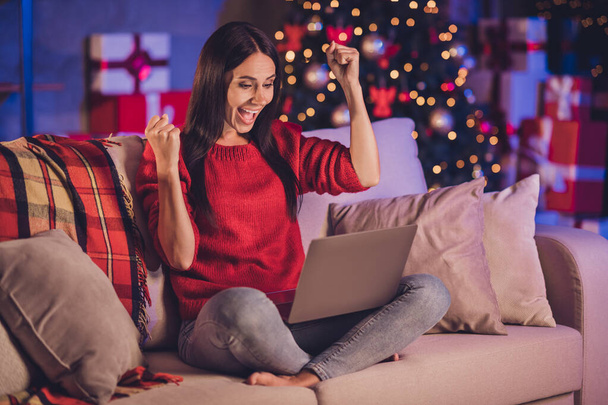 Foto retrato de la mujer sentada en el sofá en pose de loto con el ordenador portátil celebrando la victoria con los puños en el aire con decoraciones de Navidad en el interior - Foto, imagen