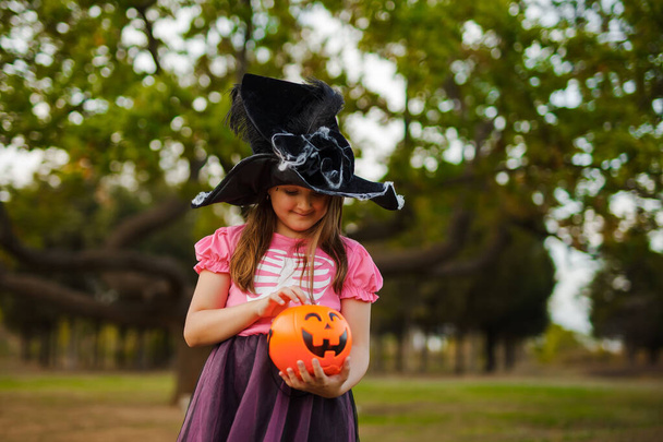 Portret van klein meisje in heks Halloween kostuum en zwarte hoed proeven snoep buiten in het bos met snoep emmer - Foto, afbeelding