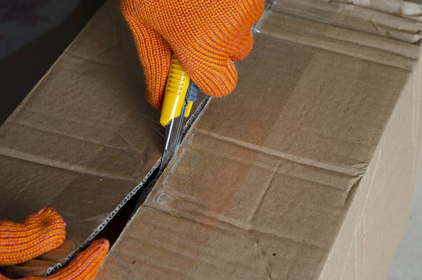 Een werknemer met beschermende handschoenen opent een kartonnen doos. Een hand met een mes snijdt de plakband op de verpakking door. Logistiek en bezorgservice. Selectieve focus. - Foto, afbeelding