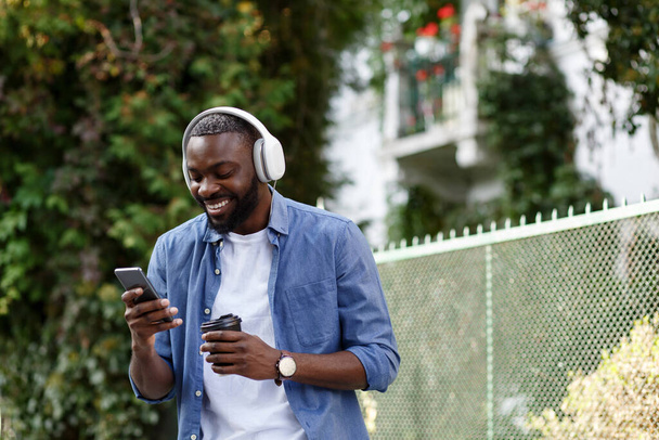 Walking in the street, relaxation, leisure. Young Afro-American man in headphones listening music on smart phone using music app. Portrait of smiling guy in earphones and mobile phone outdoors - 写真・画像