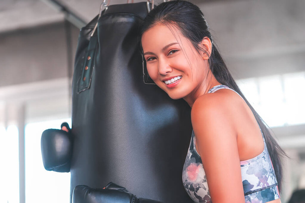 Asian woman is hugging boxing bag with smile while training in fitness gym for happy healthy lifestyle concept. - Photo, Image