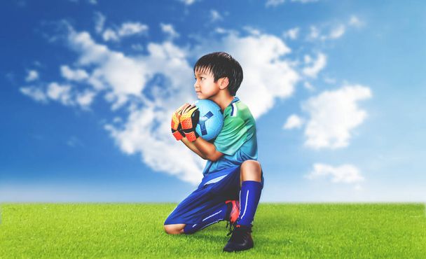Little Kid goalkeeper holding the ball on a grass field with blue sky background for success soccer dramatic concept. - Photo, Image