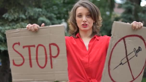Giovane donna protestante in camicia rossa tiene cartello cartello cartello di protesta con slogan Stop vaccino per la manifestazione pubblica su sfondo alberi. - Filmati, video