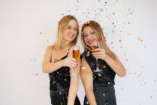 İki genç partinin fotoğrafında gülümseyen ve kadehten şampanya içen iki genç kadın beyaz arka planda izole edilmiş konfeti altında duruyorlar. - Fotoğraf, Görsel