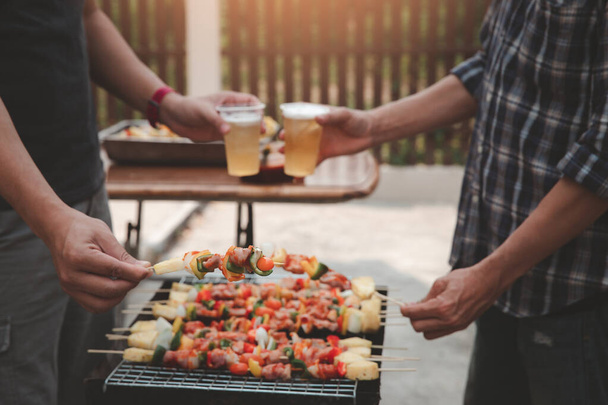 Young men cheers for a drink at a BBQ party between friends. Food, drink, people and family time concept. - Photo, Image