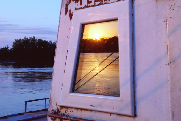 Zachód słońca nad Dunajem. Zachód słońca nad rzeką odbity w oknie starej barki transportowej w Nufaru, Rumunia. - Zdjęcie, obraz