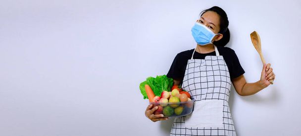 Азиатка, держащая в руках смесь овощной коробки в маске. Главная тема - "Защита и здравоохранение". Человек с копировальным пространством в помещении вид спереди с изолированным белым фоном. - Фото, изображение