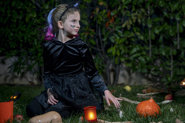 Retrato de niña en traje de bruja sentada en la noche en el parque de otoño. Niño con cubo naranja caramelo en bosque de otoño.  - Foto, Imagen