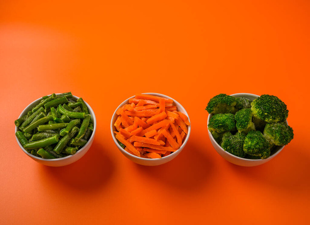 Frozen vegetables broccoli, spinach, carrot in bowls on an orange background - Photo, Image