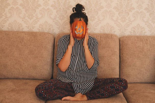 a young woman in a striped blouse sits on a sofa and holds a Halloween pumpkin near her face - 写真・画像