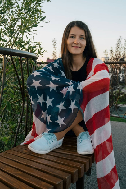 beautiful young woman covered in an American flag sits on a park bench at the evening - Foto, immagini