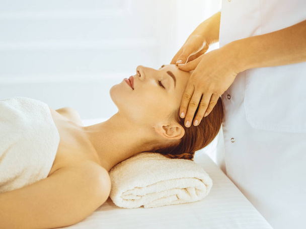 Beautiful woman enjoying facial massage with closed eyes in sunny spa center. Relaxing treatment concept in medicine - Fotó, kép