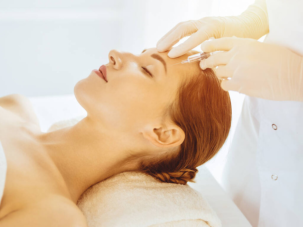Beautiful woman receiving beauty injections with closed eyes in sunny medical center. Beautician doctor hands doing beauty procedure to female face with syringe - Valokuva, kuva